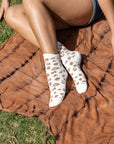 A person sits on a brown tie-dye blanket outdoors, wearing Unified Leopard Socks with a beige animal print. Only their legs and one hand are visible.