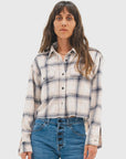 A woman with long brown hair wears the ASKK Crosby Flannel, a white and blue plaid button-up shirt, paired with high-waisted blue jeans, standing facing forward against a plain light background.