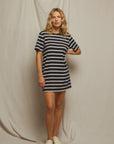 A blonde woman poses confidently with one hand on her hip, wearing the Rhett SS Tee Dress by Perfectwhitetee— a loose recycled cotton T-shirt dress— paired with white sneakers against a draped backdrop.