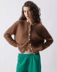 A person with wavy brown hair wears the American Vintage Vitow Button Sweater, a fuzzy alpaca wool cardigan with long sleeves and white buttons, paired with green shorts, standing against a plain light background, hands on hips.