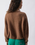 A woman with wavy brown hair faces away, wearing the American Vintage Vitow Button Sweater in brown with long sleeves and a round neck, paired with bright green shorts featuring a small embroidered design on the back pocket.