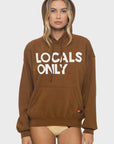A model stands against a plain white background in Aviator Nation’s “locals only - pullover hoodie relaxed” with “LOCALS ONLY” in bold white letters, hood up, paired with beige bikini bottoms.