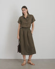 Against a plain white wall, a person wears an olive-green garment-dyed button-up shirt and the Maddie Skirt Linen by Alex Mill. They hold a small brown bag and wear brown sandals, gazing softly at the camera. The scene is minimalistic.
