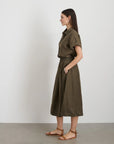 A side profile showcases the garment-dyed olive green "Maddie Skirt Linen" by Alex Mill featuring iPhone-sized pockets and a gathered waist. Long, straight hair flows as they wear brown sandals against a plain white wall on a gray floor.
