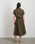 A person with long dark hair is barefoot on a light floor, facing a plain white wall, wearing the Alex Mill Maddie Skirt Linen in olive green. The skirt is knee-length with short sleeves and iPhone-sized pockets.