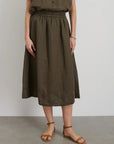 Outfitted in Alex Mill's Maddie Skirt Linen, a knee-length olive green garment with iPhone-sized pockets, paired with a matching sleeveless top. The look is completed with brown sandals and a bracelet against a plain light gray wall.