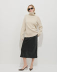 A woman stands against a white background wearing sunglasses, a beige turtleneck, brown heeled shoes, and the Alex Mill Claire Skirt—a black bias cut pull-on skirt.