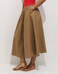 A person wearing the Alex Mill Sarah Skirt—a tan, high-waisted cotton poplin midi skirt with pockets and an invisible zipper—paired with a red sleeveless top and brown flat sandals, stands against a plain white background.