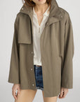 A woman layers FRAME's The Short Field Trench in olive green over a cream button-up shirt and denim shorts, standing against a plain background. Her face is partially cropped, highlighting effortless transitional style.