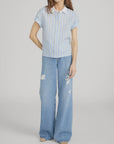 A person wears FRAME's The Boardwalk Shirt, a short-sleeve light blue and white striped linen top, paired with wide-leg light blue ripped jeans and beige shoes, standing against a plain gray background.