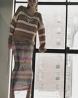 A woman stands by a large window wearing the Nirvana Sweater by Le Superbe—its chunky stripes and embellished neckline paired with a sequined skirt and open-toed heels, city buildings blurred in the background.