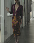 A woman with light skin stands behind a glass door, wearing a deep purple top and the Liza Skirt by Le Superbe—a gold party skirt with purple floral patterns—looking forward with her hand raised against the glass.