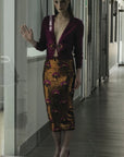 A woman with sleek, straight hair and a serious expression stands indoors behind a glass door, wearing the Le Superbe Liza Skirt—a figure-flattering sequin midi paired with a deep burgundy top—raising one hand to the glass.