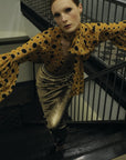 Wearing the Le Superbe Serotonin Blouse, a person stands on a staircase with arms outstretched, posing dramatically and looking up toward the camera.