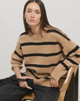 A woman with long brown hair sits on a chair, wearing the Alex Mill Striped Nottinghill Sweater and black pants. She looks slightly to the side with a neutral expression.