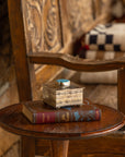 A Small Stamped Box w/ Turquoise Cross by J Alexander Rustic Silver and a hardcover book sit on a round wooden side table beside a carved bench with patterned cushions, bringing Southwestern charm to the cozy corner.