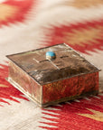 The SQ Silver Box w/ Dimples Cross by J Alexander Rustic Silver sits on a beige and red Southwestern-style geometric textile, featuring a hammered silver finish and a turquoise stone accent on its lid.