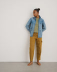 A woman stands against a white wall, wearing the Alex Mill Britt Work Jacket over an olive top, mustard pants, and tan sandals. She faces left with her hands behind her back.