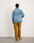 A person stands with their back to the camera wearing the Britt Work Jacket by Alex Mill, tan cargo pants, and brown sandals against a plain white wall.