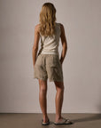 A woman with long, wavy blonde hair faces away from the camera, wearing a beige sleeveless top and James Perse Lightweight Linen Shorts in light tan with an elasticized waistband, styled casually with sandals against a plain background.