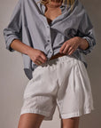 A person stands against a neutral background wearing James Perse Lightweight Linen Shorts with an elasticized waistband and a loose, light blue button-up shirt with rolled sleeves; their face is not visible.