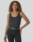 A woman with curly hair wears the FRAME Velvet Trim Tank, paired with black jeans and bracelets, standing against a light gray background and smiling softly with one hand on her hip.