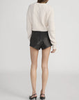 A person faces away, wearing FRAME’s The Cable Knit Cardi in white with elongated sleeves, paired with black textured high-waisted shorts and black heeled sandals against a plain light background.