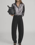 A woman stands against a plain background wearing FRAME’s The Shiny Staple Blouse in metallic gray, paired with loose black jeans, black heels, sunglasses, and carrying a large black handbag under her arm.
