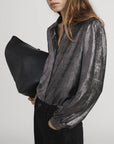 A woman in FRAME’s The Shiny Staple Blouse in metallic gray and black pants holds a large black textured bag under her arm. With long wavy brown hair and sunglasses, she stands against a simple light background.