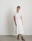 A woman smiles slightly against a white wall, wearing Alex Mill’s Maddie Skirt Linen and a matching short-sleeve 100% linen button-up shirt with iPhone-sized pockets, paired with dark crisscross-strap sandals.