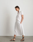 A person stands sideways by a plain white wall, wearing the Alex Mill Maddie Skirt Linen—a white midi with iPhone-sized pockets—paired with a 100% linen short-sleeved white button-up shirt and black strappy sandals, hands in pockets.