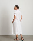 A person with short hair stands against a plain light wall, wearing the Alex Mill Maddie Skirt Linen—a white 100% linen midi skirt with iPhone-sized pockets—paired with a matching short-sleeve shirt and black strappy sandals.