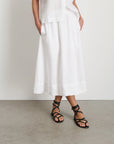 A person stands against a white wall wearing the Alex Mill Maddie Skirt Linen with iPhone-sized pockets, a white button-up top, and black strappy sandals. Concrete floor is visible; hands rest casually in the skirt’s pockets.