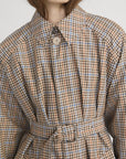 A person wears FRAME’s The Plaid Mac Coat, a belted, structured cotton blend coat with brown, blue, and beige checks, pointed collar, tailored finish, and button closure; shown from shoulders to waist with face partially out of frame.