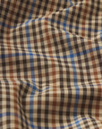 A close-up of The Plaid Mac Coat by FRAME shows its structured cotton-blend fabric in black, brown, blue, and beige checks with gentle folds and shadows on the slightly wrinkled surface.