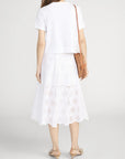 Woman faces away from the camera wearing a white short-sleeve top, the FRAME Solstice Eylet Skirt for a soft, feminine silhouette, nude flats, and carrying a brown crossbody bag.