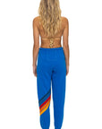 A woman with long wavy blonde hair models Aviator Nation's all seasons circle chevron women's sweatpant—blue joggers with colorful diagonal stripes—paired with a tan bikini top, standing barefoot against a white background.