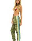 A barefoot woman poses in Aviator Nation 5 stripe women's sweatpants with blue, yellow, and green camo side stripes, paired with a colorful rainbow bikini top against a white background.