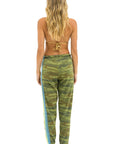 A barefoot woman with long, wavy blonde hair faces away, wearing a brown bikini top and Aviator Nation 5 Stripe Women's Sweatpant in green camo with blue and yellow side stripes for a bold, vintage-inspired style.