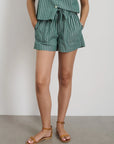 Against a white wall, a person wears Alex Mill's Montauk Pull On Shorts in Stripe with an adjustable waist and a matching top, paired with brown ankle-strap sandals. The image is cropped at the shoulders.