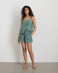 A woman with curly hair stands against a white wall wearing Alex Mill's Montauk Pull On Shorts in Stripe, a green and white vertical striped style with an adjustable waist, paired with tan sandals. Her hands are in her pockets.