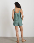 A woman with curly dark hair faces a white wall, wearing Alex Mill's green and white striped Montauk Pull On Shorts with an adjustable waist, matching top, and brown flat sandals.