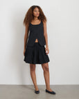 A woman with curly hair wears a sleeveless black buttoned vest, Alex Mill's true to size Penelope Pleated Skirt, and black flats, standing against a plain white wall on a light gray floor.