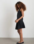 A woman with curly hair stands against a white wall, wearing the Alex Mill Penelope Pleated Skirt in black, true to size, paired with a sleeveless top and black ballet flats. She faces sideways with her head slightly tilted down.
