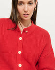 A woman with straight brown hair wears the Alex Mill Nico Chunky Cardigan in bright red cotton knit with large cream Corozo buttons and a round neckline. She pairs it with gold hoop earrings and gazes slightly past the camera, expression neutral.