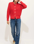 A woman wears the Alex Mill Nico Chunky Cardigan in bright red with Corozo buttons, paired with blue jeans and beige shoes, standing against a white background. She touches her hair and looks slightly downward.