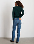 A woman with curly hair, wearing the Alex Mill Alice Polo Cashmere in dark green, blue jeans, and black shoes stands with her back to the camera facing a white wall in a minimal indoor setting.