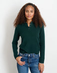 A woman with curly brown hair wears the Alex Mill Alice Polo Cashmere sweater in dark green and blue jeans, standing against a plain white background with one hand in her pocket.