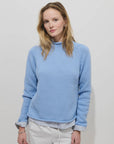 A woman with light brown hair wears the Alex Mill Eastport Rollneck in Cotton—a light blue sweater—over a striped shirt and white pants, standing against a plain white background, looking at the camera with a slight smile.