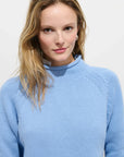 A woman with blonde hair wears the Alex Mill Eastport Rollneck in Cotton, a light blue 100% cotton sweater, and looks at the camera with a slight smile against a plain white background.